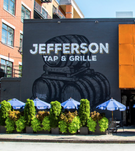 The Jefferson Tap & Grille Storefront. It's a black building with a mural of stacked kegs. In front of the building are green bushes along with tables and chairs that are shaded by blue umbrellas.